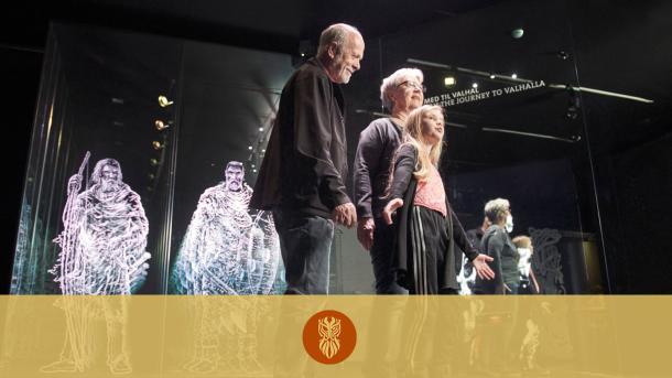 Flere ældre og yngre museumsgæster daner en kæde i den interaktive udstilling Rejsen til Valhal. I baggrunden ses lysende Vikinger.